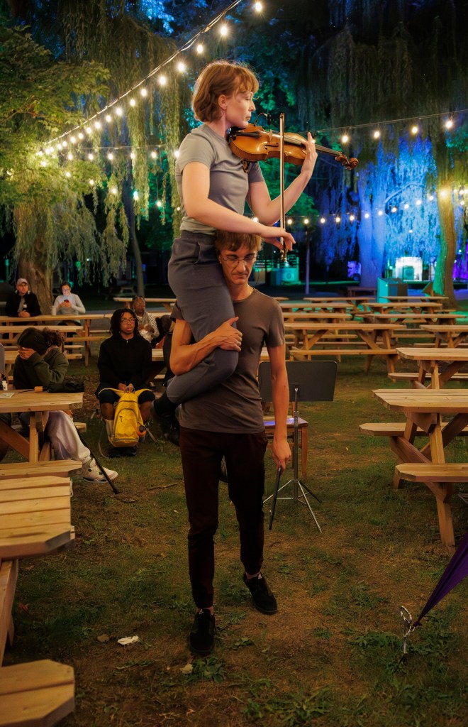 Dance artist Chloe Hart sits on the shoulder of her collaborator playing the violin over his head. He is walking forward. It is evening and the light is dim.