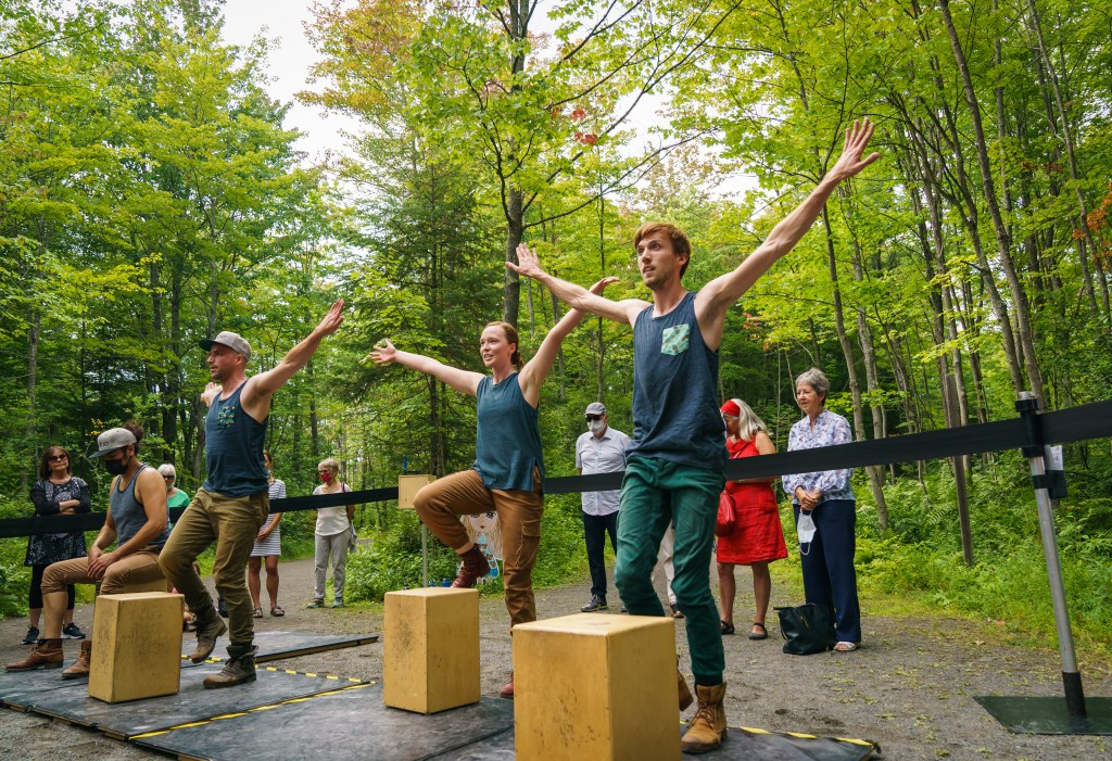 Performing Zeugma Danse's Cube, Chloe Hart is in the background, dressed in a blue tank top and brown pants, slightly behind fellow dancer Louis Roy. Both dancers have their arm stretched in a V and prepare to step onto a cajón, a box-shaped instrument. They are in a lush forest.