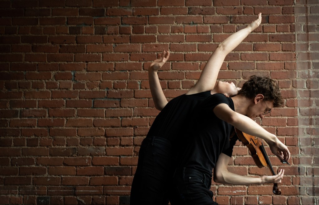 In front of a red brick wall, a dancer, Chloe Hart, and violinist, Julien Oberson are positioned back to back. Julien leans forward, rounding his back while playing, and Chloe arches backwards with her arm outstretched.
