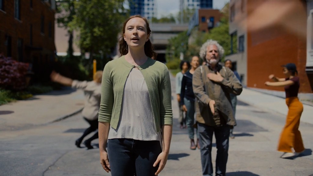 Chloe Hart, dressed in a green cardigan and faded black jeans, sings while dancers move around her. They are standing in the street on a bright sunny day.